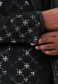 Fekete dzseki, textúrált anyagból készült, flitterekkel díszítve, ami virágmintát alkot, fekete gombokkal a mandzsettán.
