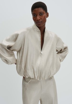 Vrouw met kort haar die een losse beige jas met rits draagt en bijpassende broek, staand met handen in haar zij tegen een effen achtergrond.