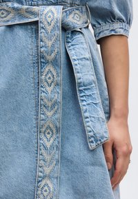 Φόρεμα από ανοιχτό μπλε τζιν με ζώνη κεντημένη με σχέδια, διακοσμημένη με γεωμετρικά μοτίβα στο εμπρός μέρος και στα μανίκια.
