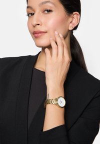 Gouden horloge met een ronde witte wijzerplaat, schakelband en duidelijke markeringen; model draagt een zwart blazer en eenvoudige gouden stud oorbellen.