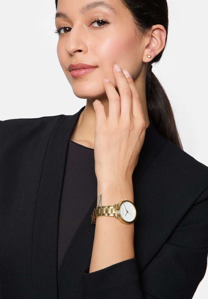 Gouden horloge met een ronde witte wijzerplaat, schakelband en duidelijke markeringen; model draagt een zwart blazer en eenvoudige gouden stud oorbellen.