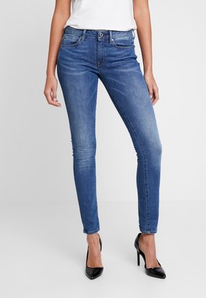 Femme portant un jean en denim bleu ajusté et des chaussures à talons noirs, debout devant un fond clair uni.