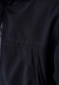 Giacca nera con zip, caratterizzata da una texture liscia, con logo ricamato "PROJECT X PARIS" e un emblema stilizzato "RP" sul petto.