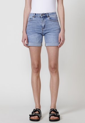 Shorts in denim blu chiaro con orli arrotolati, dotati di chiusura frontale con bottone, tasche posteriori e un lavaggio leggermente sbiadito. Abbinati a sandali neri.
