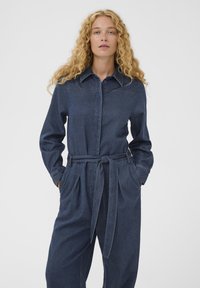 Ženska s kodrasto blond lasmi, oblečena v temno modro džins jumpsuit z dolgimi rokavi in zavezanim pasom, roke v žepih, nevtralna ozadje.