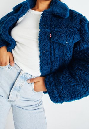 Person iført en blå fluffy jakke over en hvid ribbet skjorte og lyseblå højtaljede jeans med hænderne i lommerne.