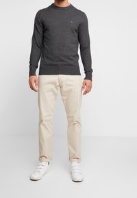 Pull gris foncé à manches longues et col rond, associé à un pantalon beige et des baskets blanches. Le matériau semble doux et texturé.