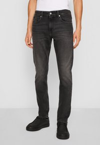 Jeans de mezclilla negra con corte slim, que presentan un diseño clásico de cinco bolsillos, un cierre de cremallera y un botón de metal en la cintura. Textura suave.