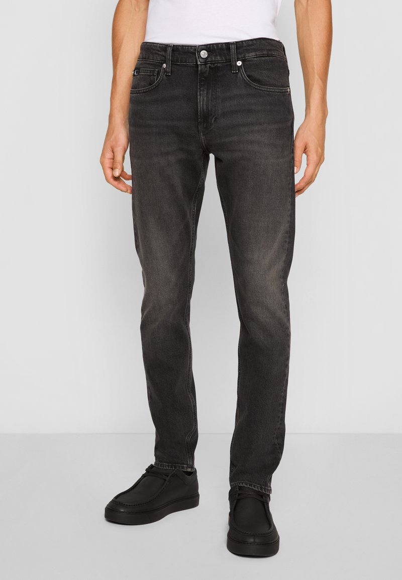 Jeans de mezclilla negra con corte slim, que presentan un diseño clásico de cinco bolsillos, un cierre de cremallera y un botón de metal en la cintura. Textura suave.