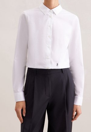 Kvinde iført en tætsiddende hvid skjorte med knapper og en lille sort rosebroderi over sorte højtaljede plisserede bukser.