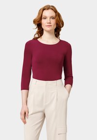 Bodysuit à manches longues bordeaux associé à un pantalon ample crème présentant une poche avant et des plis pour un design structuré.