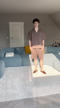 Camisa de manga curta castanha e calções a condizer feitos de um tecido leve, combinados com sapatos castanhos, em pé sobre um tapete de cor clara.