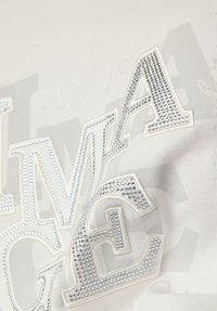 Nahaufnahme eines weißen Stoffes mit aufgenähten Buchstaben "LMA GE", verziert mit kleinen glänzenden silbernen Nieten in einem dekorativen Muster.