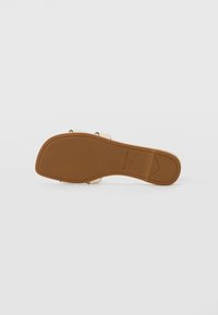 Semelle de sandale plate beige avec une large bride beige. Présente un léger talon, une texture en caoutchouc et un détail de logo embossé sur la semelle.
