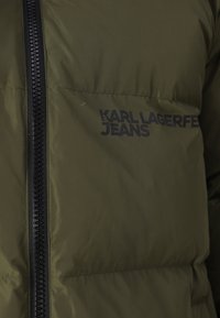 Alyvuogių spalvos pūkinė striukė su juodu užtrauktuku. Yra „KARL LAGERFELD JEANS“ prekės ženklo užrašas juoda spalva. Lygi tekstūra, su siuvinėta dizaino forma.