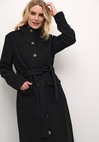 Manteau noir en tissu doux, avec un col haut, des boutons dorés, une ceinture à nouer, et deux poches plaquées à l'avant.