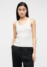 Νεαρή γυναίκα με ίσια μαύρα μαλλιά, φορώντας λευκό αμάνικο ριμπ τοπ και μαύρο ψηλόμεσο παντελόνι, στέκεται μπροστά σε μονόχρωμο φόντο.