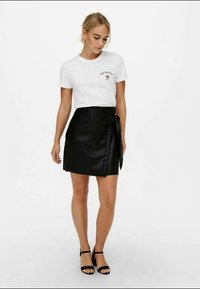 T-shirt bianco con stampa "GO NUTS", abbinato a una gonna avvolgente nera in pelle sintetica con legatura laterale e finitura liscia. Sandali neri completano il look.