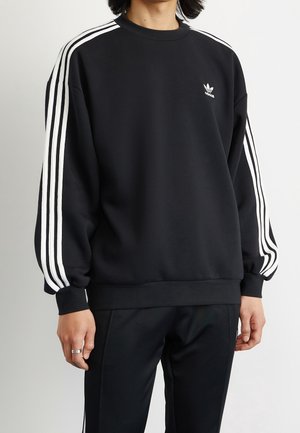 Personne portant un sweat-shirt Adidas noir avec trois bandes blanches sur les manches et le logo Adidas sur la poitrine, associé à un pantalon noir.