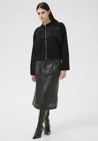 Frau, die einen schwarzen, fransenbesetzten Reißverschluss-Pullover, einen schwarzen, perforierten Leder-Midirock und schwarze, Absatzstiefel trägt, steht vor einem schlichten Hintergrund.