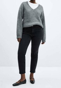 Personne portant un pull gris à col en V, une chemise blanche en dessous, un jean noir taille haute et des chaussures plates en cuir verni noir, debout sur un fond blanc.