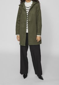 Olivgrön kappa med huva, tryckknappsstängning och långa ärmar, bärs över en vit randig tröja och svarta byxor. Svarta spetsiga skor.