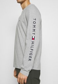 Šedé tričko s dlouhými rukávy vyrobené z bavlny, s textem "TOMMY HILFIGER" v námořnické modři podél rukávu a malým logem na prsou.