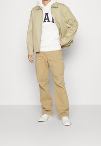 Giacca beige leggera sopra un hoodie bianco con testo, abbinata a pantaloni cargo beige e sneaker bianche. Outfit casual a strati.