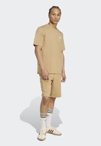 Korte mouwen t-shirt en shorts set in tan katoen, met een klein wit logo. Gekleed met beige schoenen en witte sokken met bruine strepen.