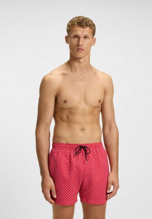 Red swim shorts with a white geometric pattern, elastic waistband, and black drawstring. The fabric appears lightweight and smooth.