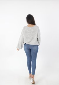 Lichtgrijze oversized gebreide trui met brede mouwen, gedragen over blauwe skinny jeans. Het model staat met de rug naar de camera en toont een relaxte pasvorm.