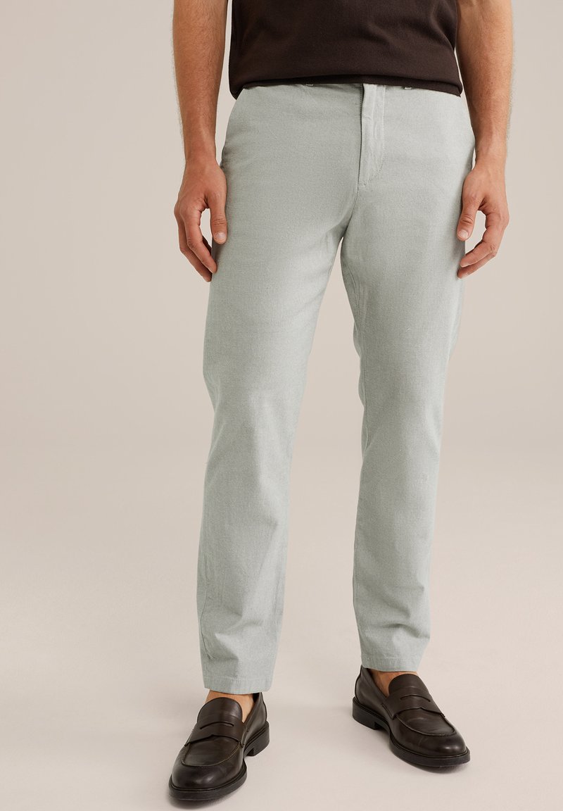 Svetlo zelene, teksturirane hlače s slim fit krojem in ravnim sprednjim delom. Sovpadajo s temnimi loafer čevlji, kar poudarja formalno, a hkrati sproščeno zasnovo.