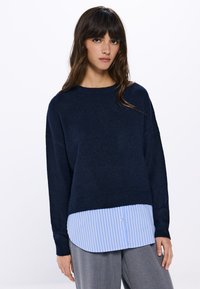 Marineblauwe gebreide trui met lange mouwen over een lichtblauw en wit gestreept overhemd met kraag. Zachte textuur, relaxte pasvorm en gelaagd ontwerp.