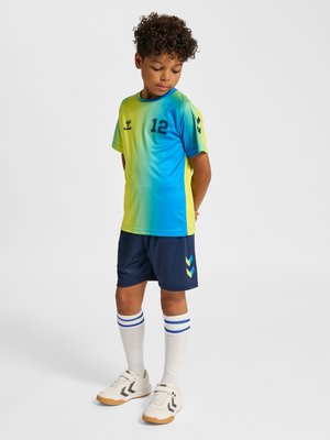 Jongen met krullend haar die een blauwgroen sportshirt met kleurverloop en nummer 12 draagt, marineblauwe korte broek, witte sokken met blauwe strepen en witte sneakers.