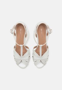 Weiße Riemchensandalen mit glatter Textur, Knoten-Design vorne und dünnem Knöchelriemen. Ausgestattet mit einem niedrigen Absatz und beigem Innenfutter.