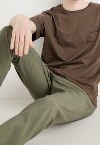 T-shirt in cotone marrone con colletto a girocollo e grafica di teschio. Abbinato a pantaloni aderenti verde oliva con tasche laterali e superficie morbida.