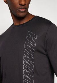 Modèle masculin portant un t-shirt noir à manches longues avec un grand texte vertical réfléchissant "HUMMEL" sur la poitrine, visible du cou jusqu'au milieu du torse.