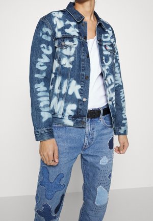 Veste en jean avec texte blanc style graffiti sur un jean bleu clair présentant plusieurs patchs cousus bleu foncé et blanc.