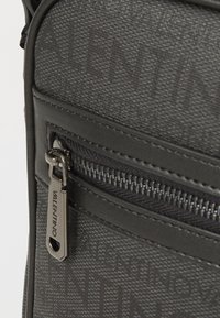 Sac en bandoulière noir avec un motif texturé, une poche zippée munie d'un tirette de marque et des coutures raffinées.