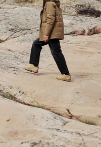 Personne portant une veste beige, un pantalon noir et des chaussures beiges marchant en montée sur un terrain rocheux.