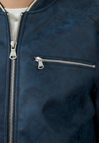 Veste bomber bleu marine au toucher lisse, dotée d'une fermeture éclair frontale argentée et d'une poche poitrine zippée. Accentuée d'un col clair.