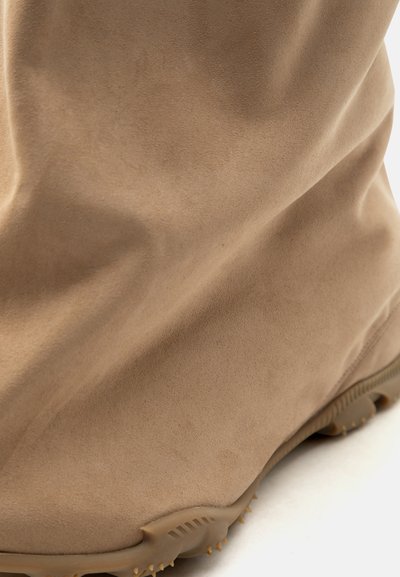 Κοντινή λήψη μπεζ μποτίνι από σουέντ με στρογγυλεμένη μύτη και υφασμένο καουτσούκ πέλμα σε λευκό φόντο.