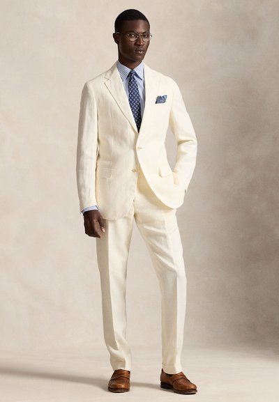 Kreminės spalvos lininis kostiumas su dviem sagomis, atvartinėmis atlapais, kišenėmis ant siūlės; derinamas su šviesiai mėlyna dryžuota marškiniais ir raštuota kaklaraiščiu. Rudi odiniai batai.