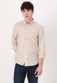 Camisa de botones a rayas beige con cuello. Mangas largas arremangadas hasta los codos. Pequeño logo en el pecho. Combinada con unos jeans azul oscuro.