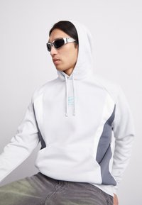 Valge kapuutsiga sviiter hallide paneelidega, millel on ees tasku ja helesinine logo. Mudel kannab peegeldavaid prille.