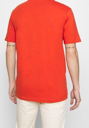 Homme portant un t-shirt uni orange vif à manches courtes et un pantalon clair, vu de dos avec des tatouages de fil barbelé sur les deux bras.