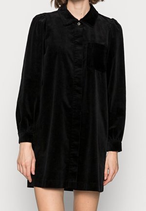 Zwart corduroy overhemdjurk met lange mouwen, knoopsluiting aan de voorkant, borstzak en licht opgeblazen schouders, gedragen door een staande persoon.