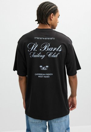 Camiseta oversize negra con texto blanco: "Club de Navegación de St. Barts", coordenadas geográficas y "Indias Occidentales Francesas del Caribe" en la parte posterior.