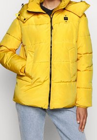 Gele pufferjas met een ritssluiting aan de voorkant, zijzakken en een afneembare capuchon. Gladde stof met horizontale doorgestikte patronen.