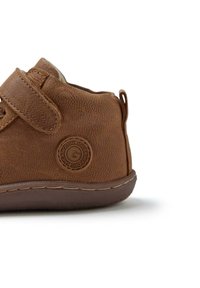 Zapato de cuero marrón con acabado texturizado, correa de Velcro, forma redondeada y un acento de logo circular en el lateral. Suela de goma con detalles de costura.